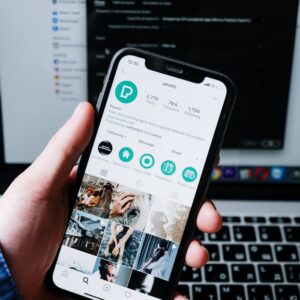 Hand holding smartphone displaying social media app with laptop in the background.